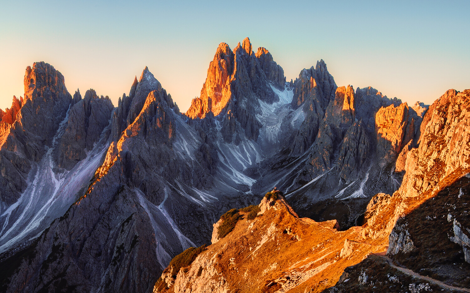Jagged alpine peaks bathed in golden sunrise light with snow patches and dramatic shadows
