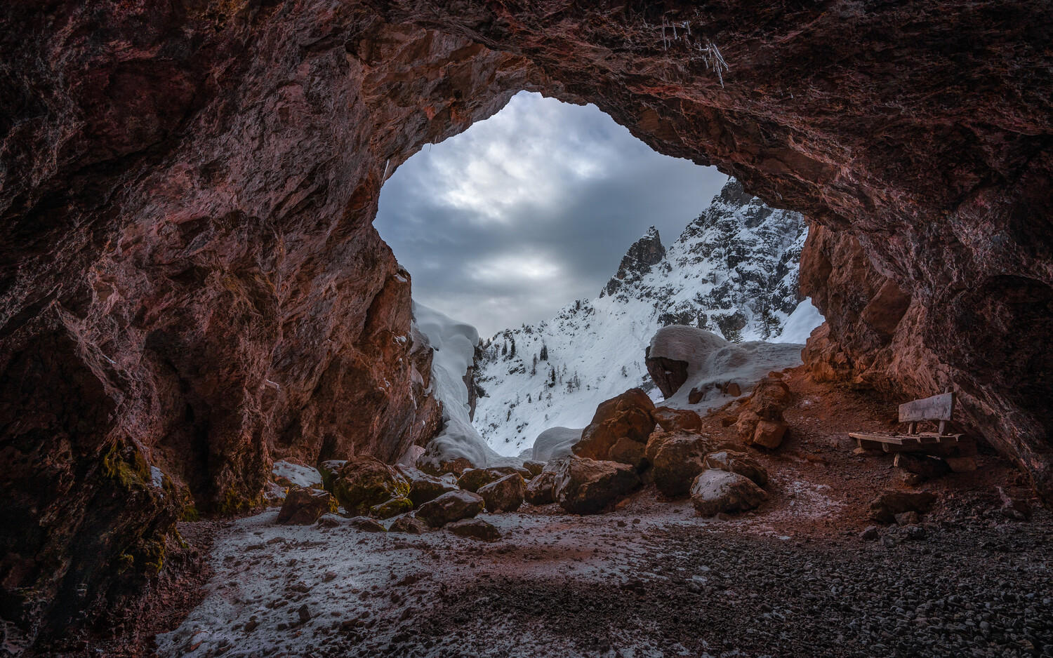 View from inside a reddish rock cave through an arched opening to snow-dusted mountain peaks under an overcast sky