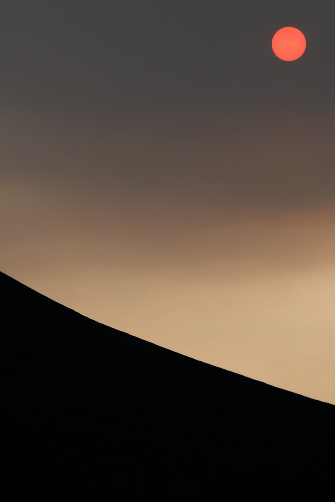 Dark silhouetted ridge curving across the lower frame beneath a hazy sky with a red-orange sun in the upper right.