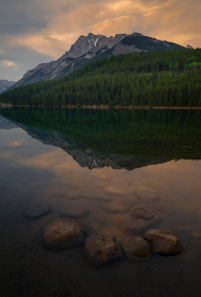 Mountain reflected in still lake at dusk with forested shoreline and rocks in foreground