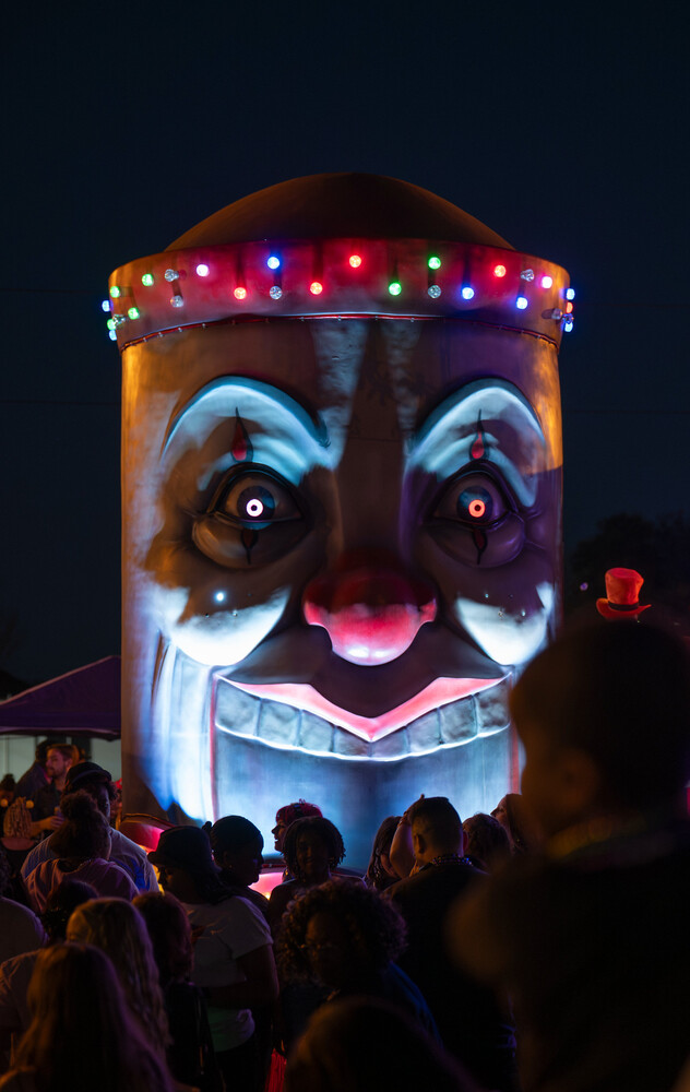 Glowing face sculpture with wide eyes and pink lips, topped with colorful string lights, towering over a nighttime crowd.