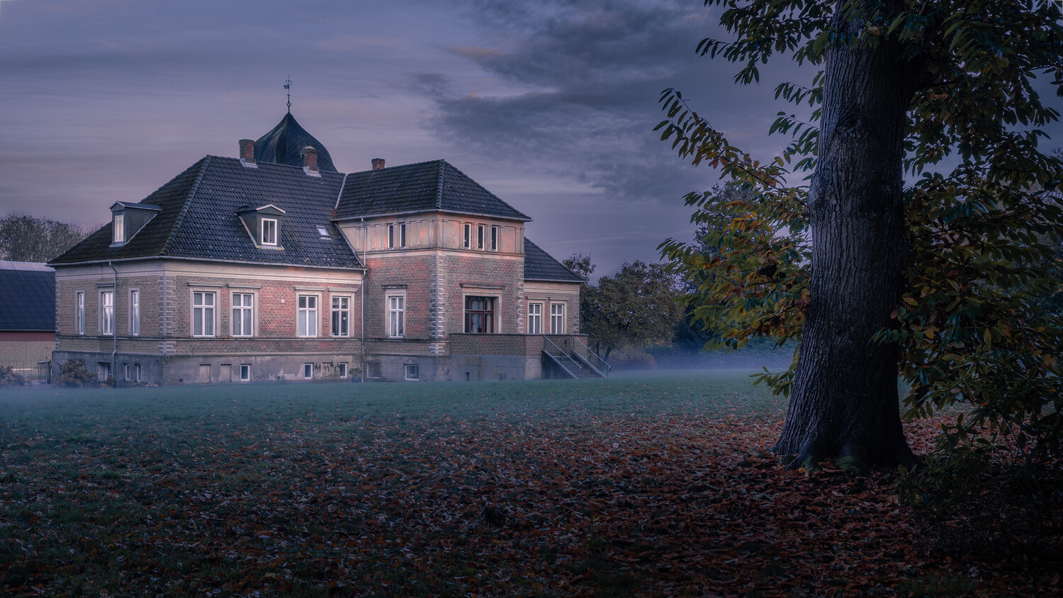 Pink mansion with mansard roof at dusk, surrounded by autumn foliage and mist