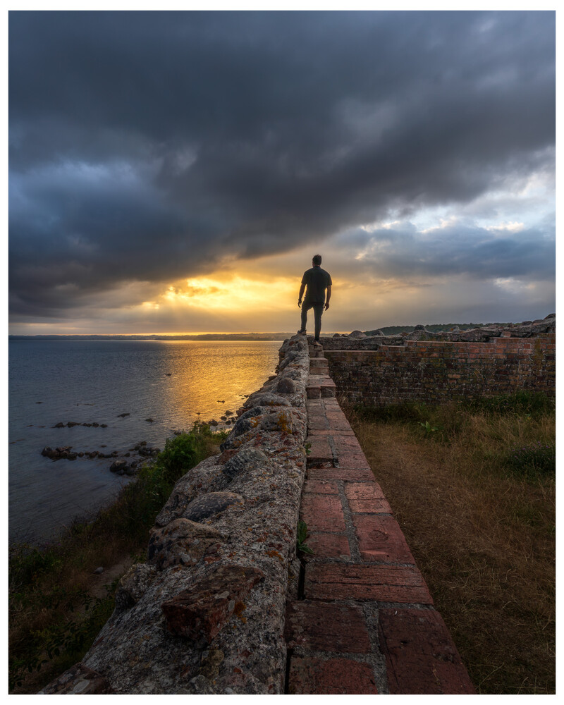 Silhouetted figure standing on a stone wall at sunset over water