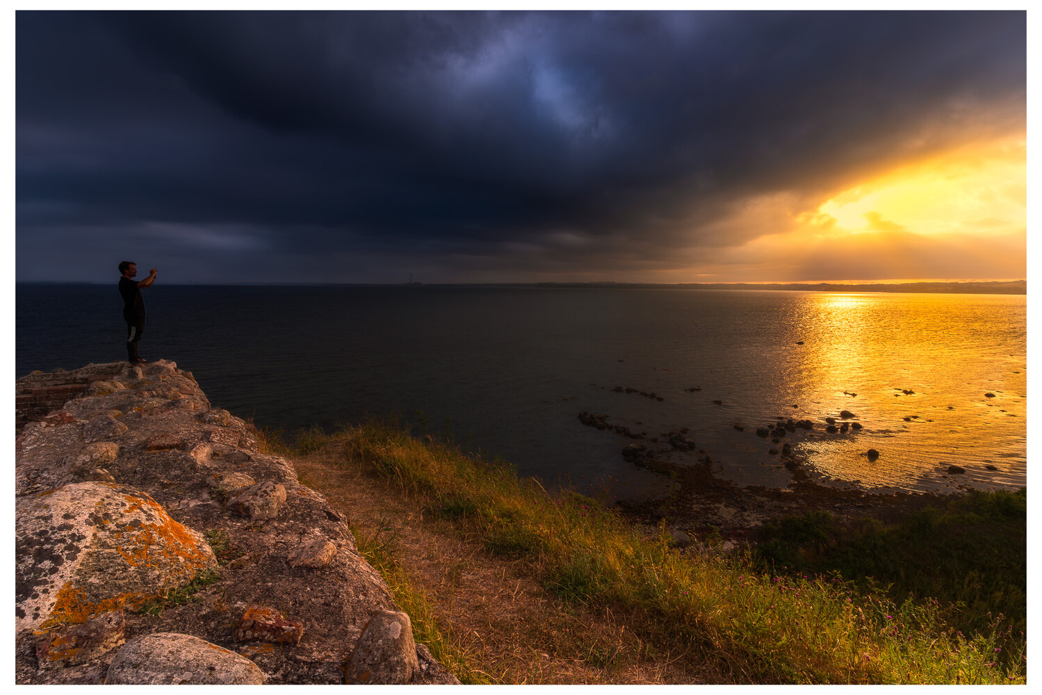 Solitary figure standing on rocky outcrop overlooking calm water at sunset