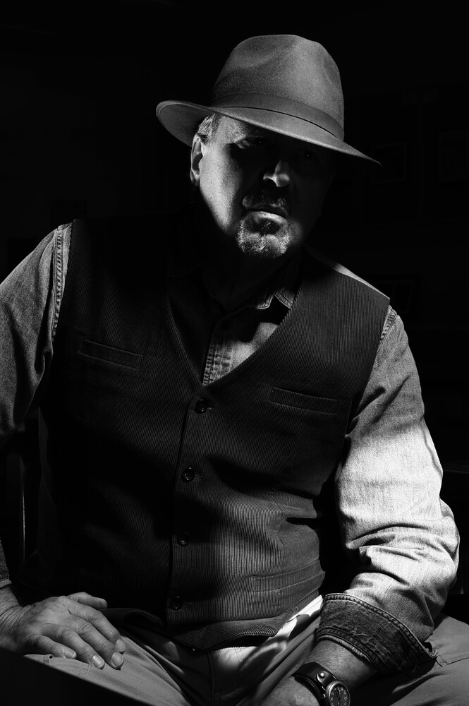 Black-and-white low-key portrait of a bearded man in a fedora and dark vest, lit dramatically against a black background.