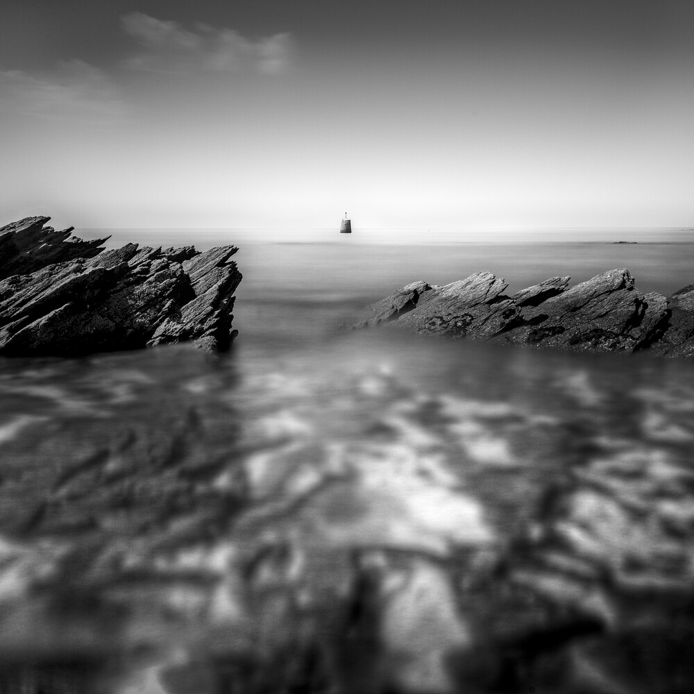 Rocky coastline with smooth water and distant lighthouse in monochrome