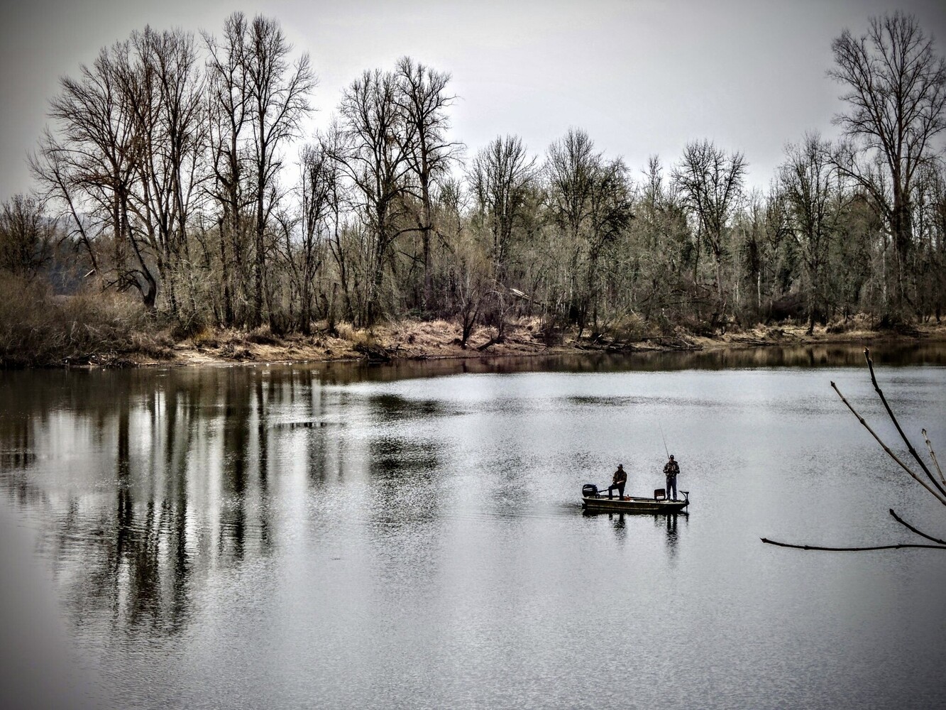 Two fishermen in a small boat on a calm river lined with bare trees