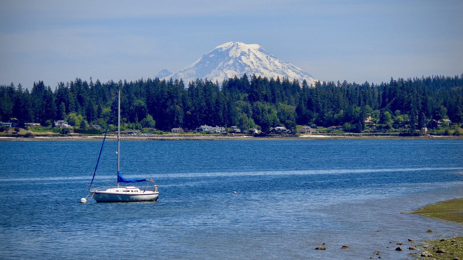 Sailboat anchored in calm blue water with snow-capped mountain and forested shoreline in background