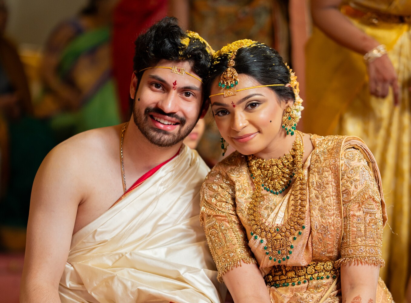 Smiling couple in traditional Indian wedding attire, woman in gold temple jewelry and flowers, man in cream silk dhoti