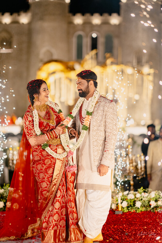 Smiling couple in red and gold bridal attire and a cream sherwani, both wearing garlands, at a night wedding celebration.