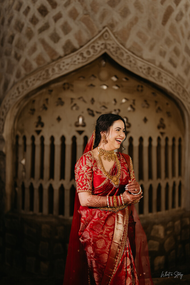 Smiling woman in a red and gold embroidered bridal saree and heavy gold jewelry, posed before an ornate carved stone arch.