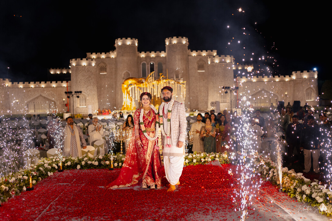 A bride in a red and gold embroidered lehenga and a groom in a cream sherwani stand smiling on a red rose petal aisle at night, flanked by cold sparkler fountains.