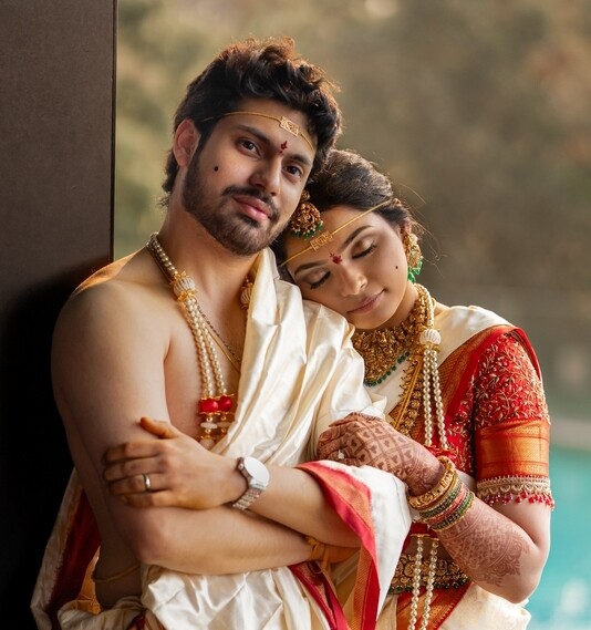 Couple in traditional wedding attire embracing, the woman resting her head on the man's shoulder with eyes closed, against a blurred green background.