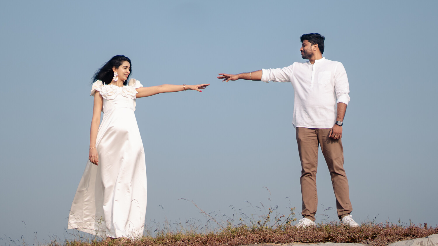 Woman in a white ruffled gown and man in a white shirt and khaki trousers stretch their arms toward each other against a pale blue sky on a grassy hilltop.