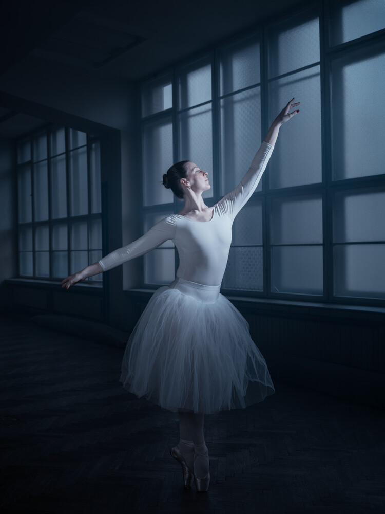 Ballet dancer en pointe in arabesque pose by large windows in moody blue-toned studio