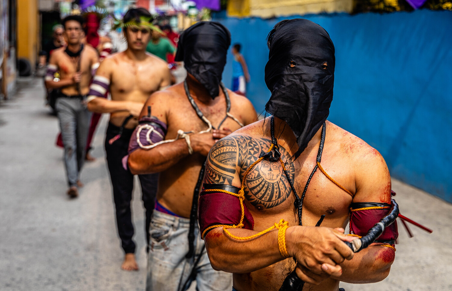 Shirtless performers with body paint and black face coverings walk in a street procession