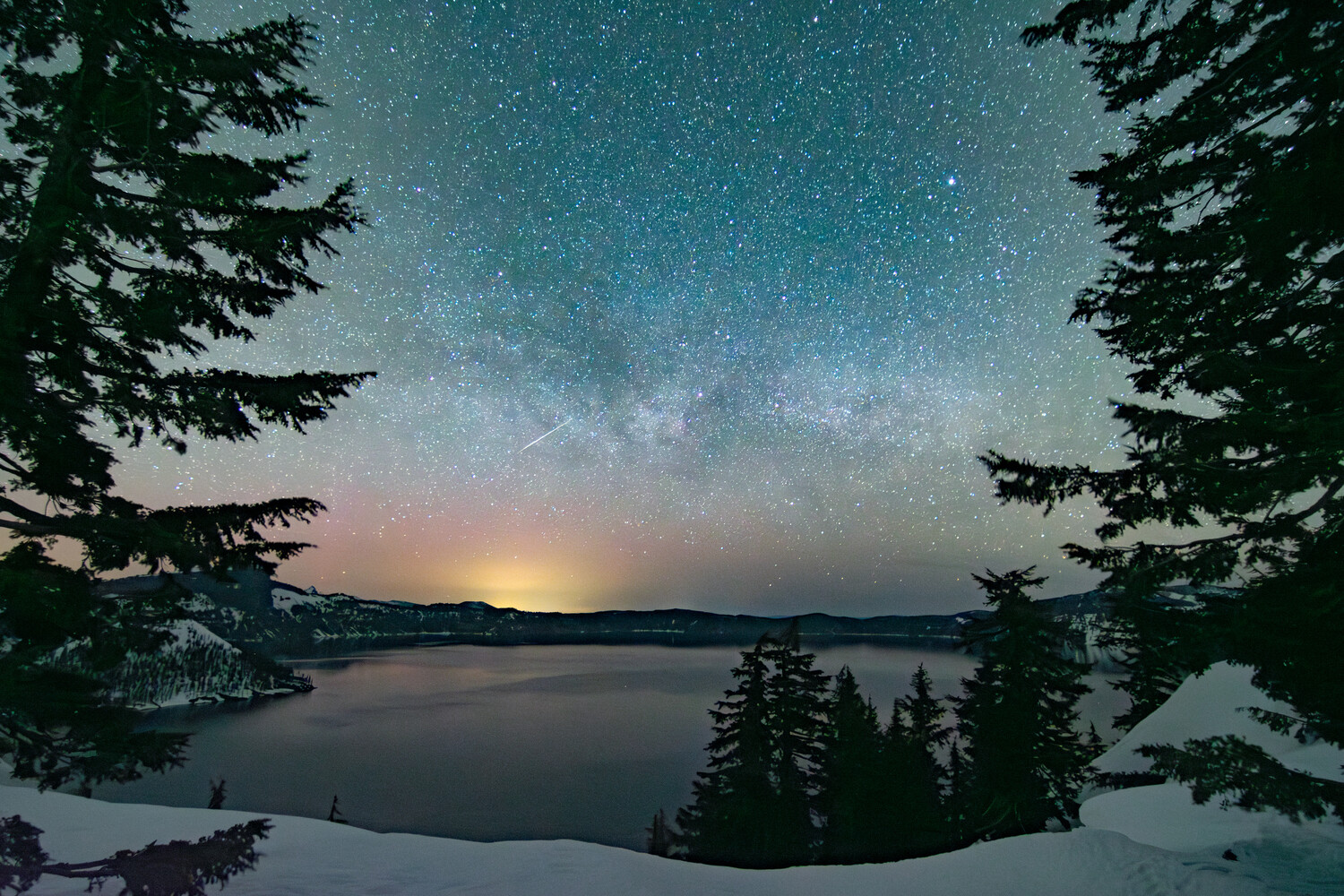 Milky Way galaxy arches over a snow-dusted alpine lake at twilight, framed by evergreen trees