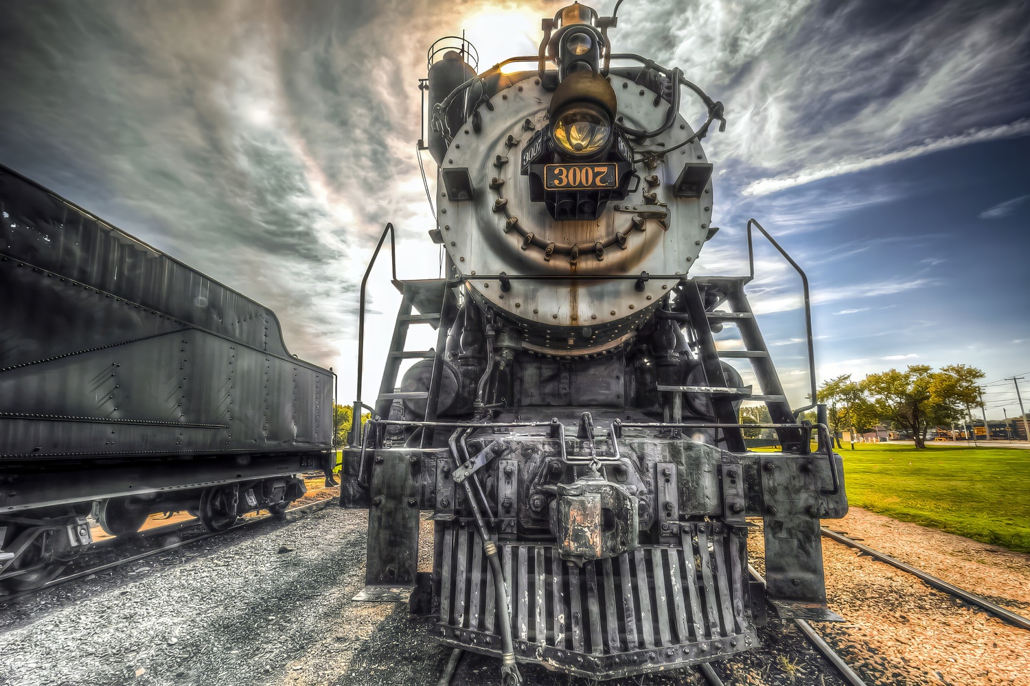 Front view of a black steam locomotive numbered 3002 on display tracks under a dramatic cloudy sky
