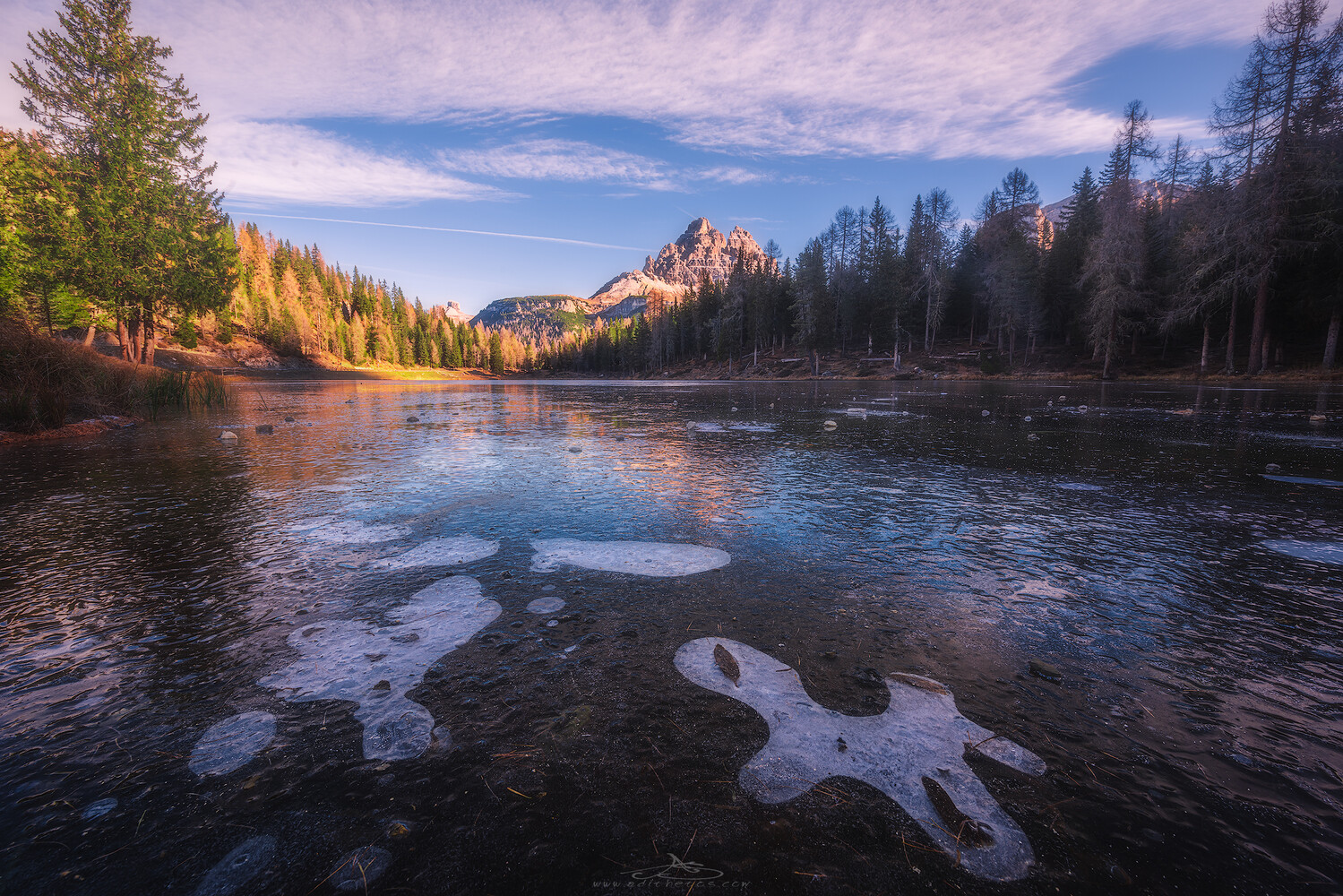 Alpine lake with thin ice sheets in the foreground, conifers lining the shore and a jagged mountain peak under a blue sky.
