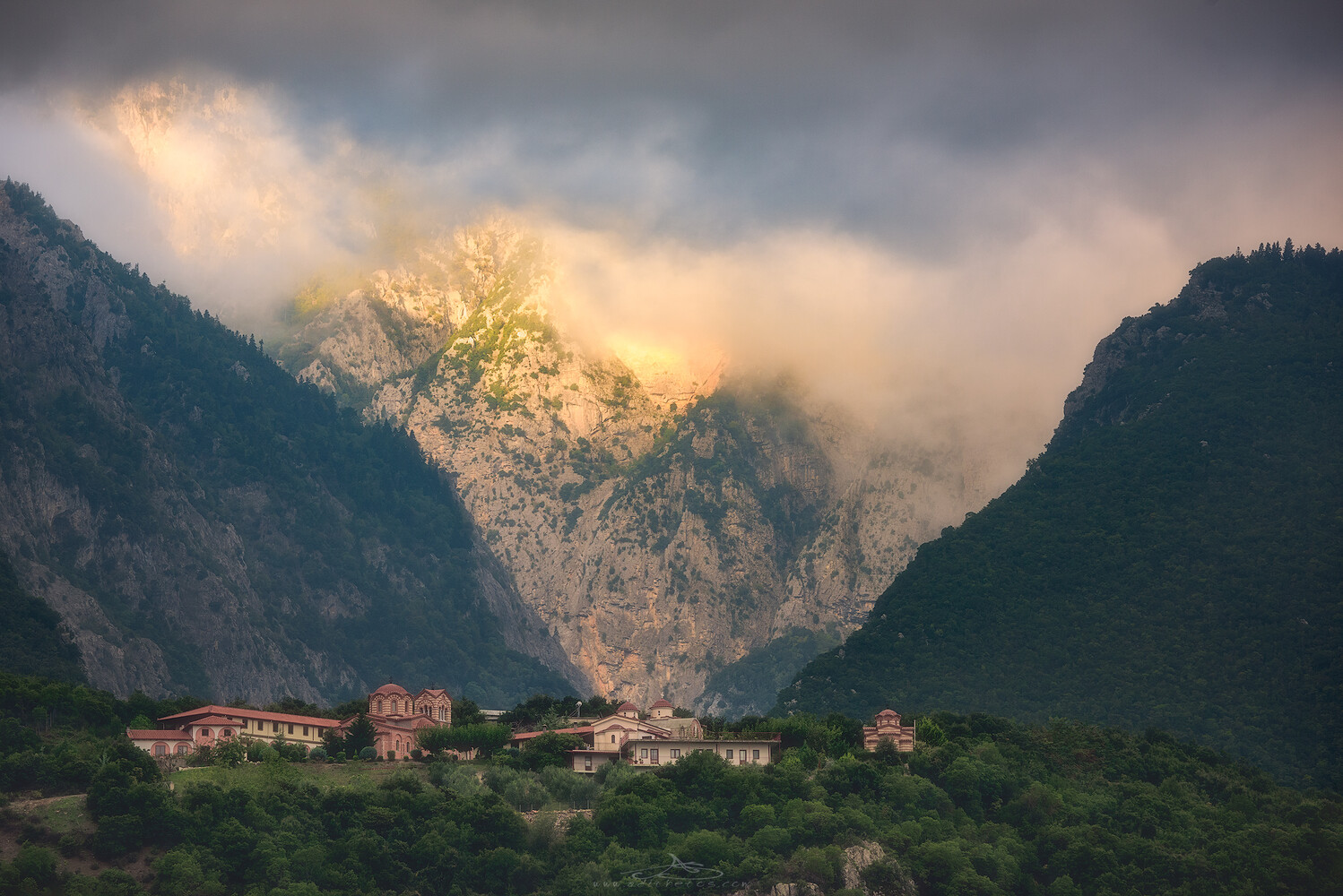 Misty mountain peaks lit by warm sunlight breaking through clouds, with a hillside village of terracotta-roofed buildings nestled among dense green trees below.
