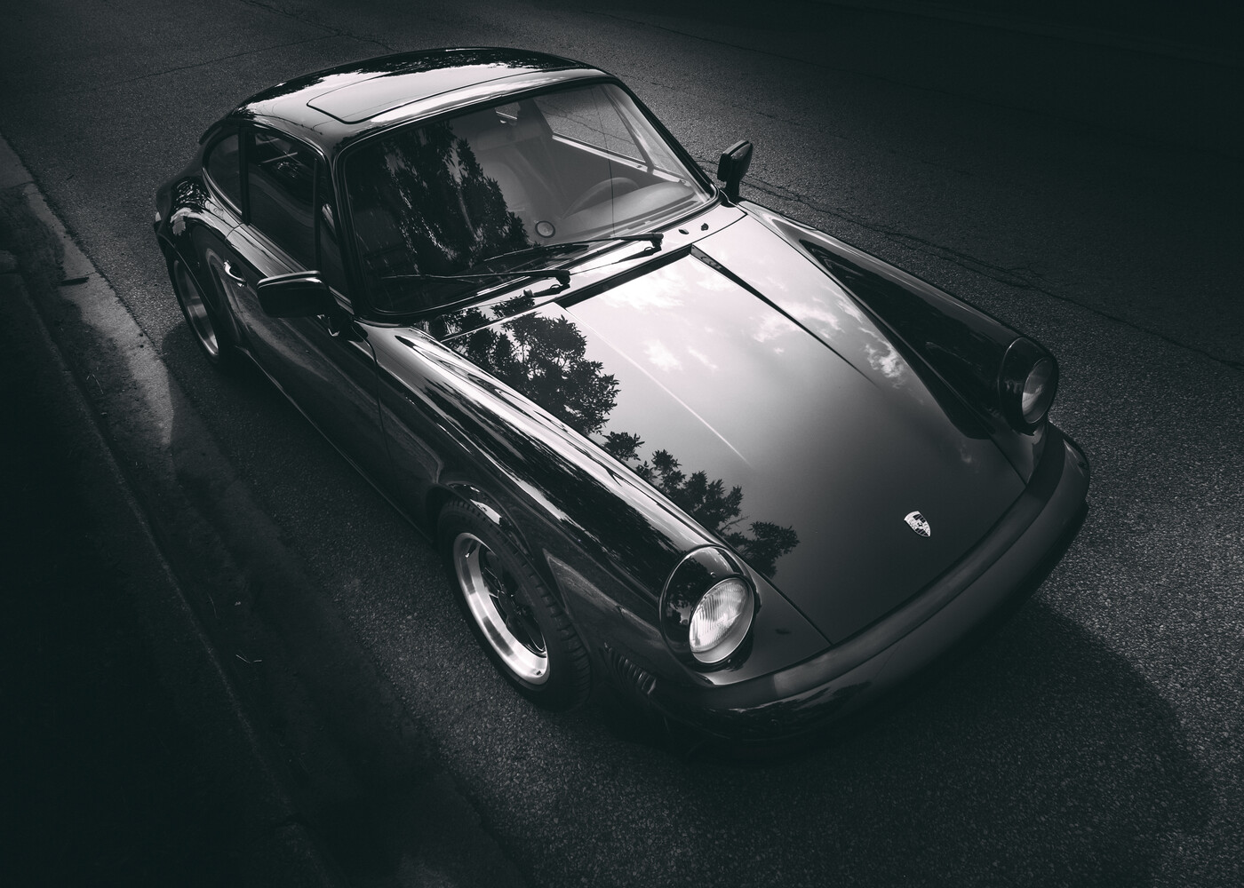 Monochrome photo of a classic Porsche 911 coupe viewed from above, with tree reflections across its hood.