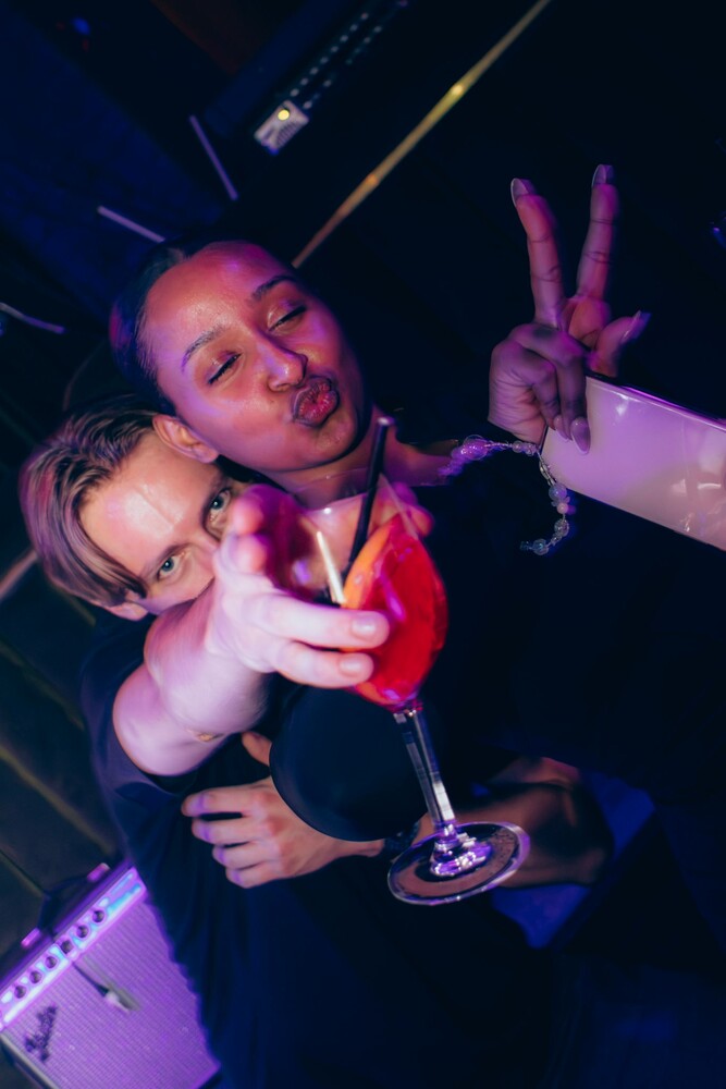 Person holding a cocktail making a peace sign under purple and pink stage lighting