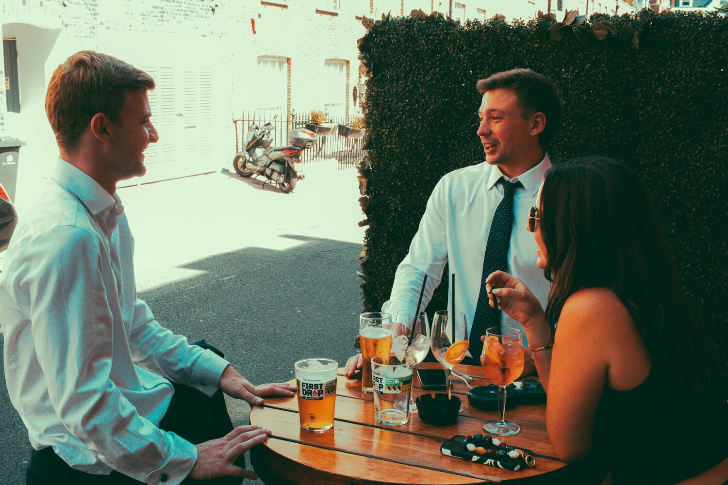 Three people in business casual attire conversing at an outdoor table with drinks and cigarettes