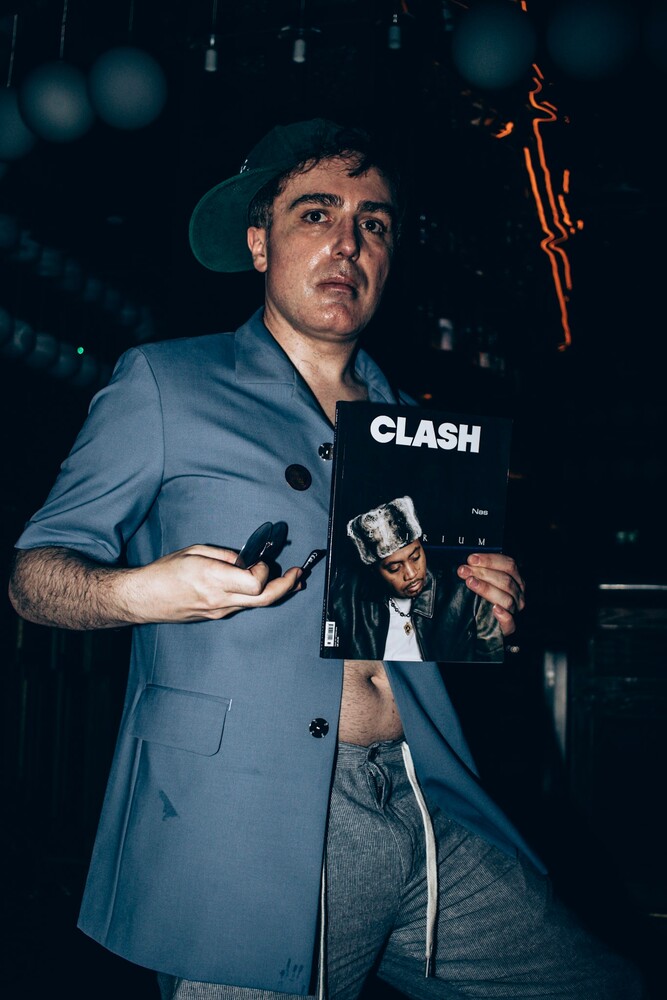 Man in light blue blazer holding microphone against dark backdrop with text reading 'CLASH'