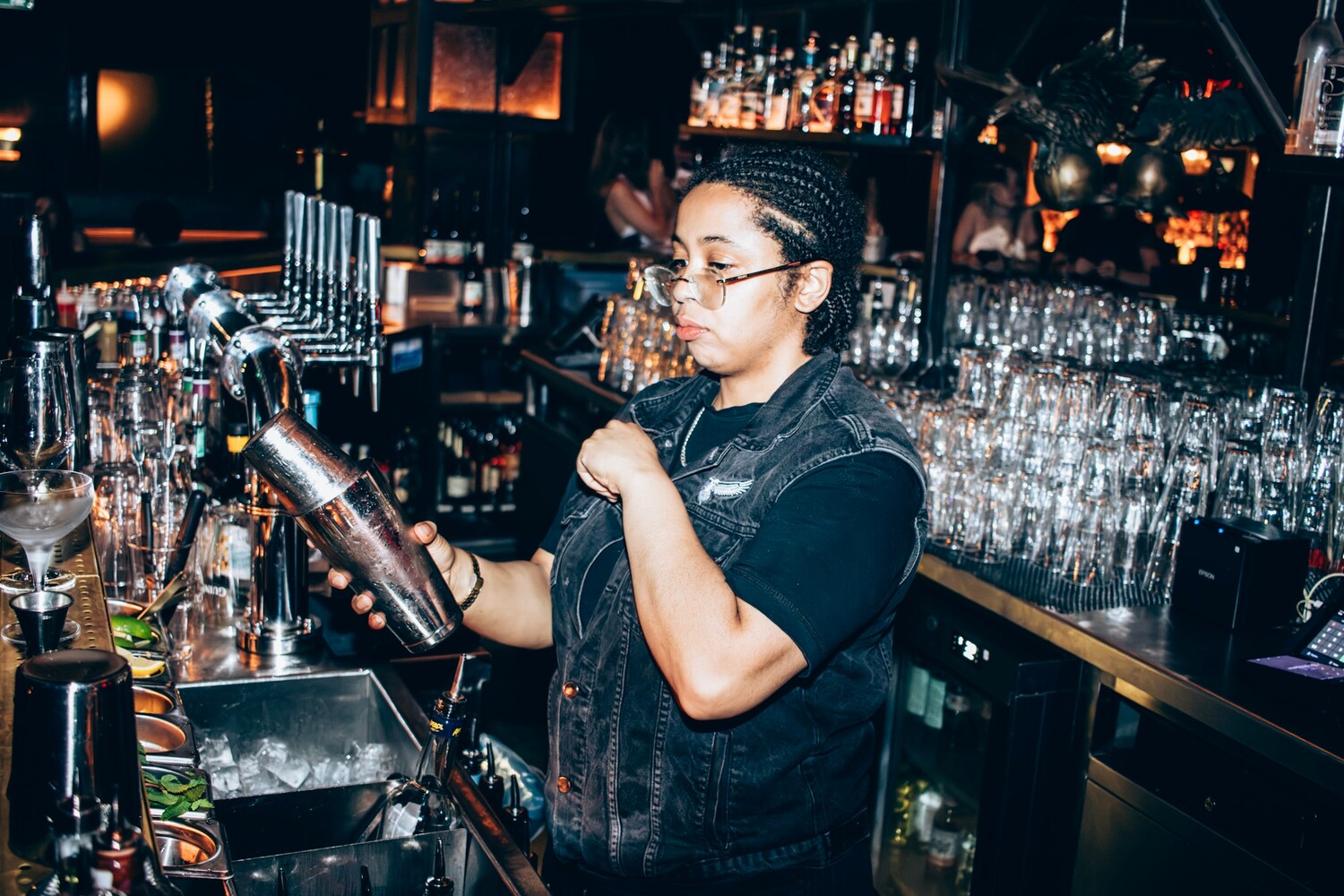 Bartender in glasses and denim vest pouring a cocktail behind a well-lit bar counter