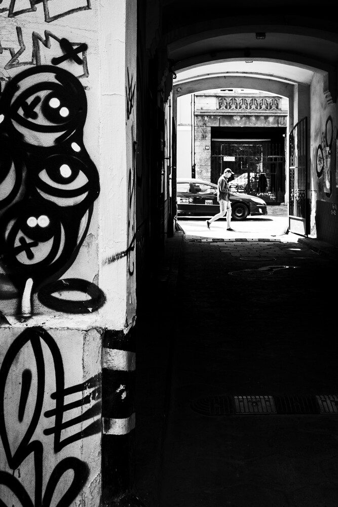Black and white street photograph showing a narrow passageway with decorative street art on the left and a distant storefront visible through the archway
