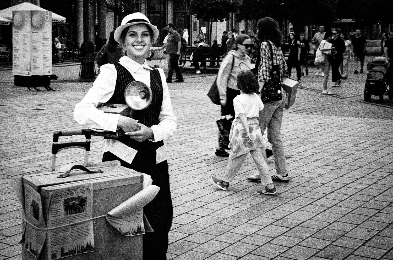 Street performer in vintage-style clothing poses with a suitcase prop in a crowded public square