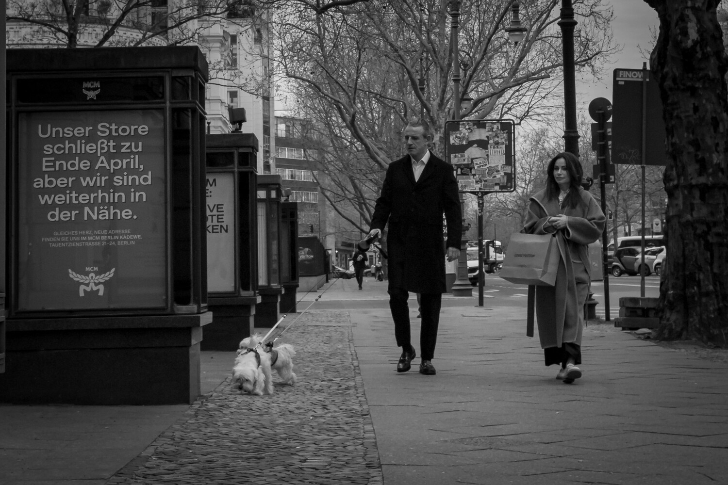 Black and white street scene with pedestrians walking past a storefront sign in German