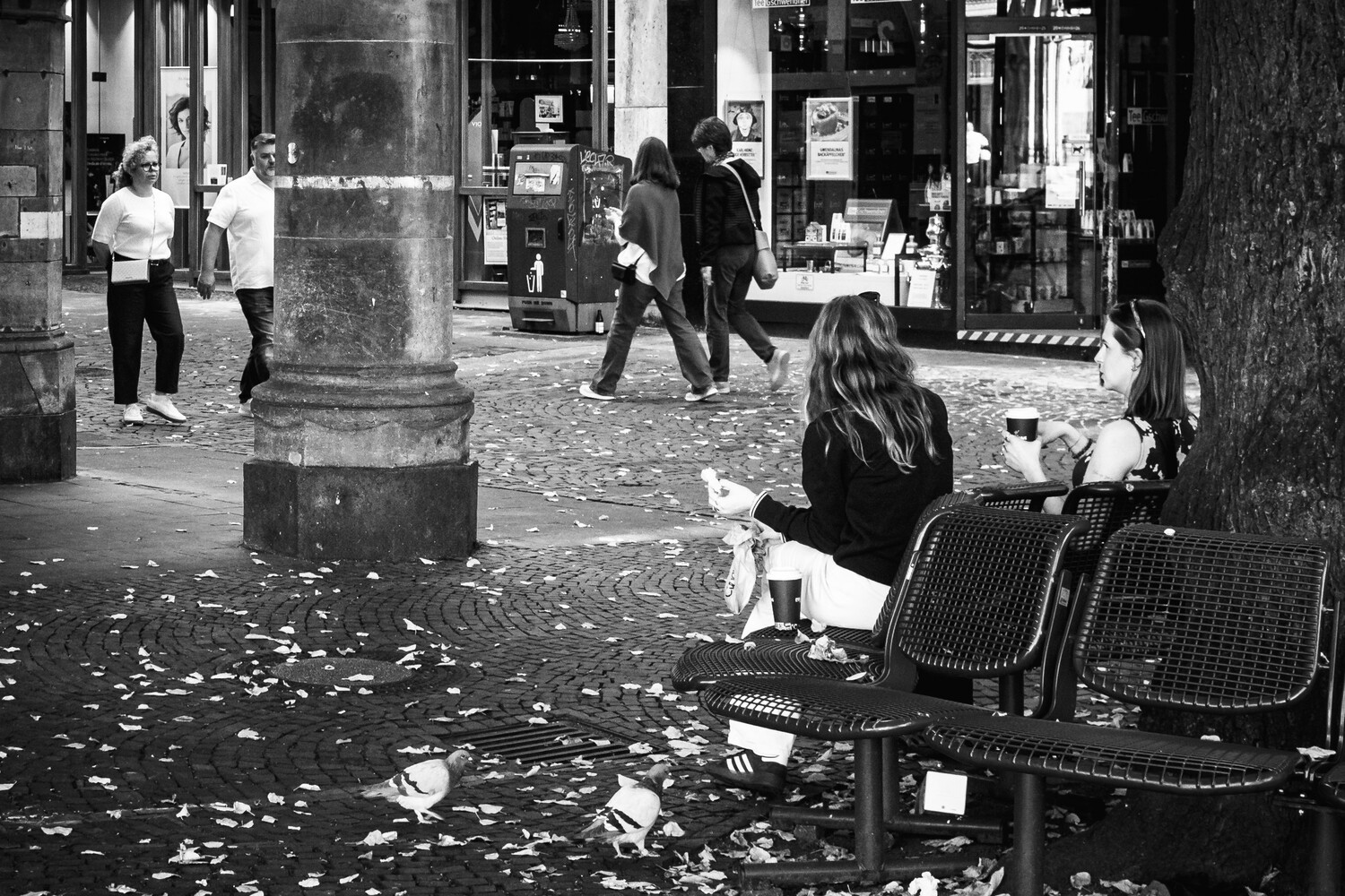 Woman seated on street-side chairs with long dark hair, photographed from behind in black and white
