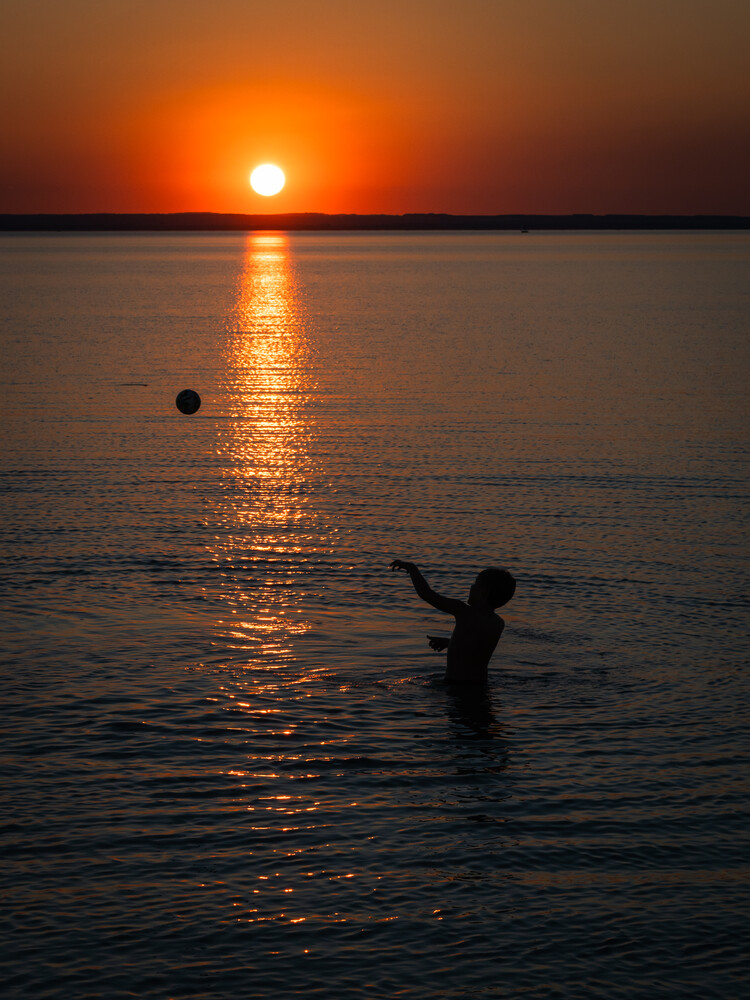 Silhouetted figure wading in calm water during vibrant orange sunset