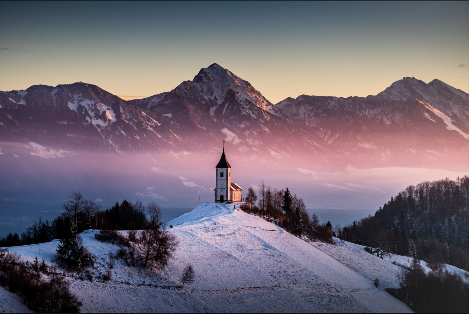 Church with steeple on snowy hilltop at dawn with alpine mountains and valley fog
