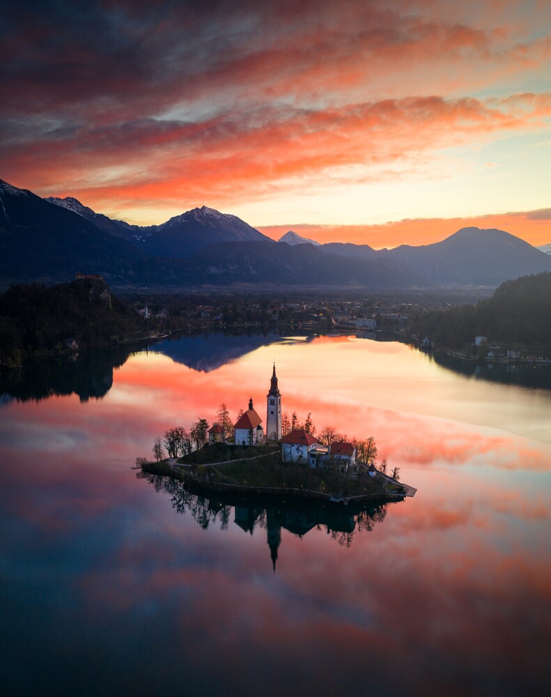 Island church with illuminated spire reflected in glassy lake at sunrise