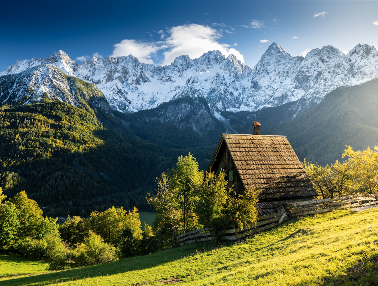 Wooden alpine cabin with golden roof nestled in sunlit meadow beneath snow-capped mountain peaks