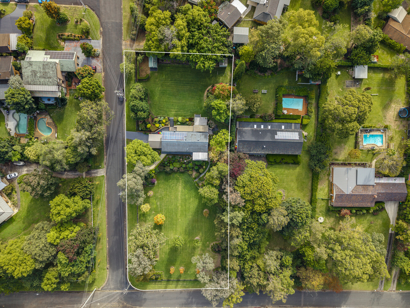Aerial view of a residential property with manicured lawn, swimming pool, and dark-roofed house surrounded by mature trees