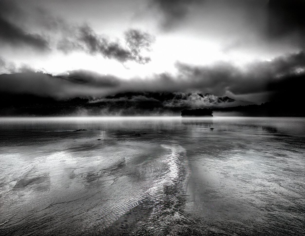 Dramatic monochrome seascape with rippled sand, distant mountains, and storm clouds