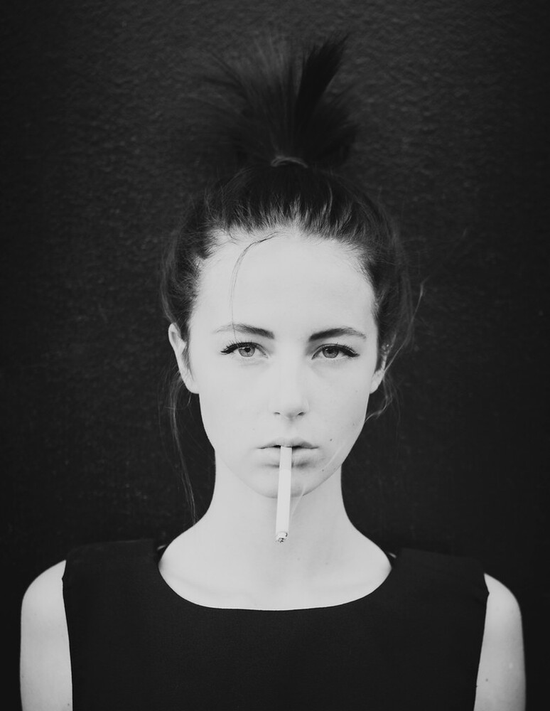 Black and white portrait of a woman with a cigarette between her lips, hair pulled up, against a dark textured backdrop
