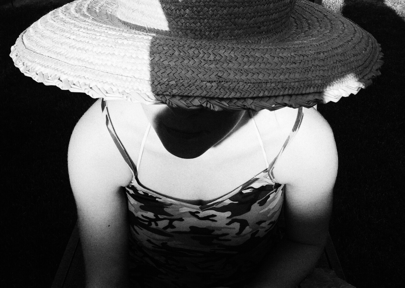Black-and-white photo of a person in a wide straw hat and camouflage camisole, face hidden under the brim in hard sunlight
