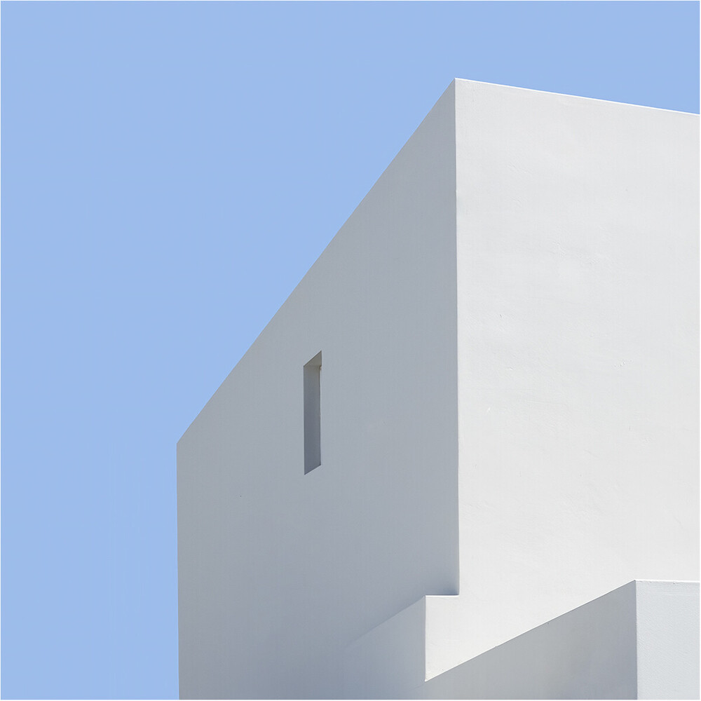Sunlit white building corner with a small recessed window against a clear pale blue sky