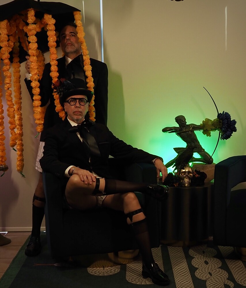 Person in formal dark suit seated against backdrop with orange marigold garland decoration