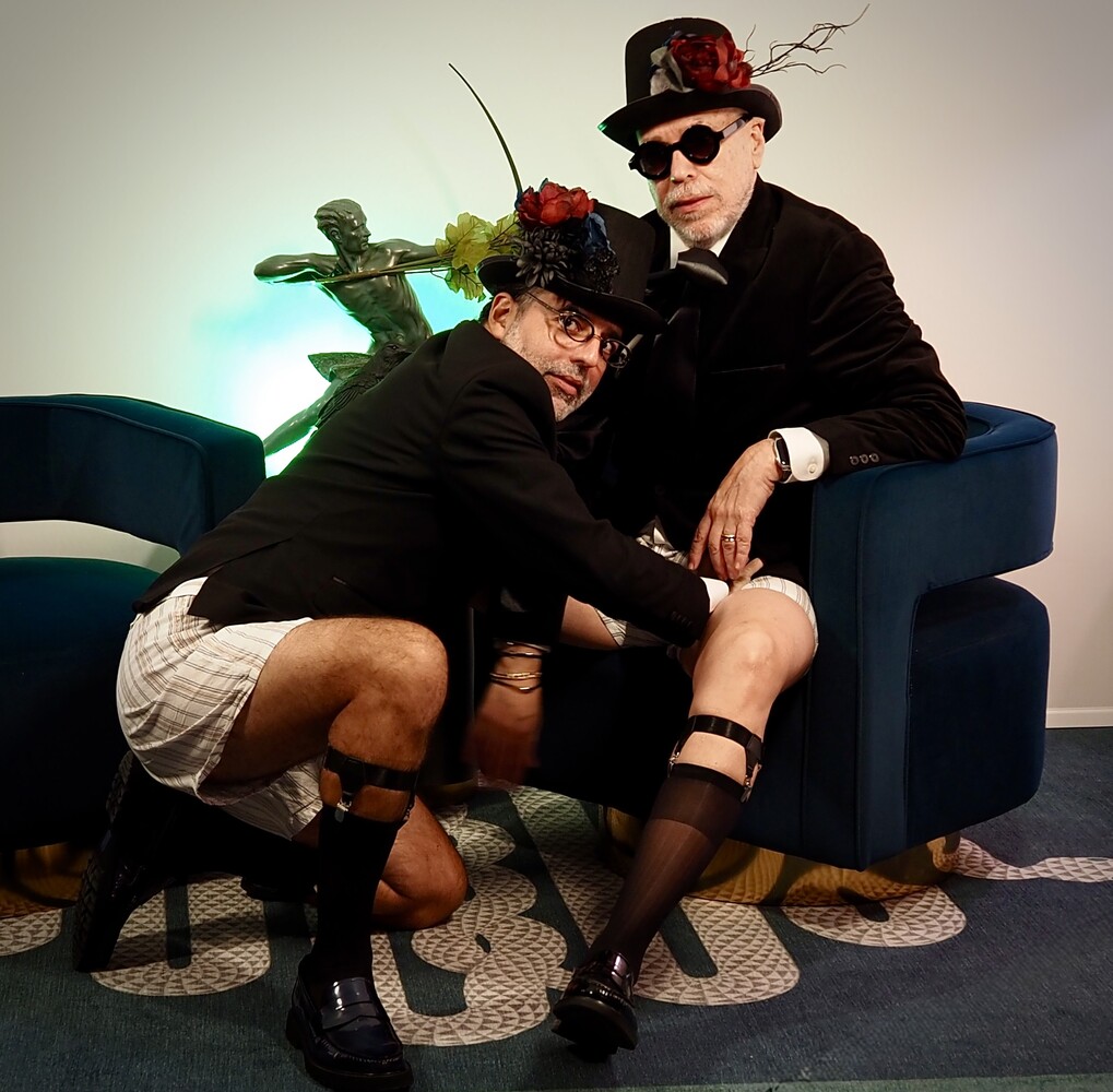 Two people in black suits and hats pose playfully on a blue couch with decorative plant