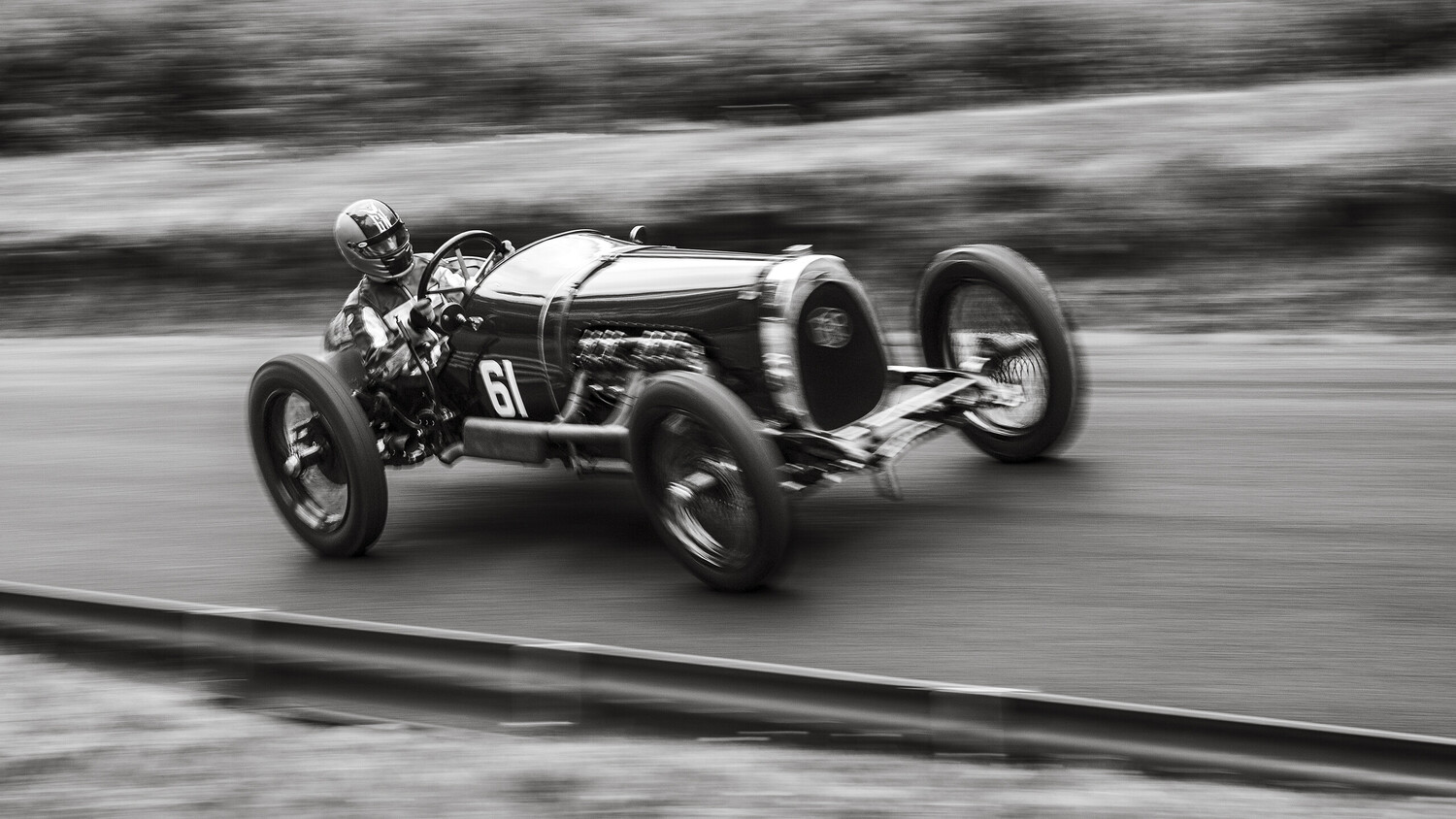 Black-and-white panning shot of a vintage racing car numbered 61, with a helmeted driver, speeding along a road past a guardrail.