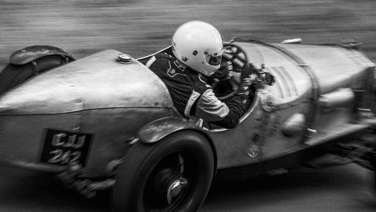 Black and white panning shot of a driver in a white helmet piloting a vintage silver race car at speed.