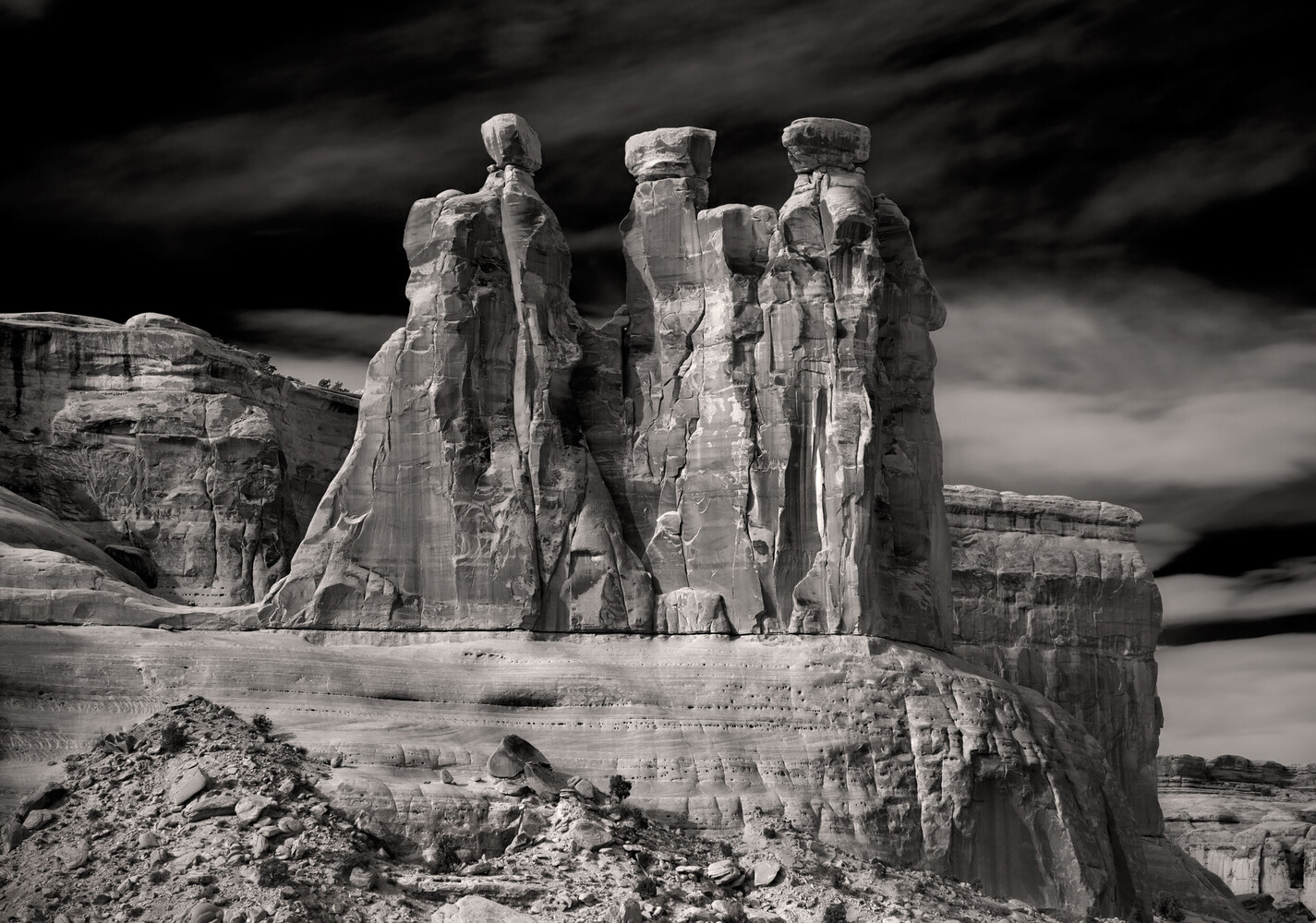 Black and white view of three tall sandstone spires rising from a massive rock pedestal beneath a dramatic, cloud-filled sky