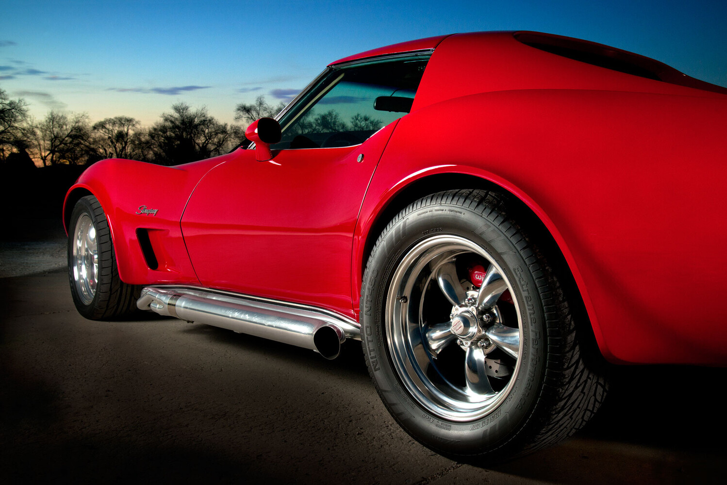 Low-angle view of a glossy red classic Corvette with chrome side exhaust and polished wheels, against a dusk sky and dark trees.