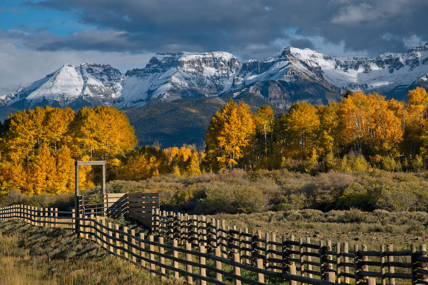 Snow-dusted mountain range rising above golden aspen trees and a split-rail fence in a meadow under a cloudy autumn sky.