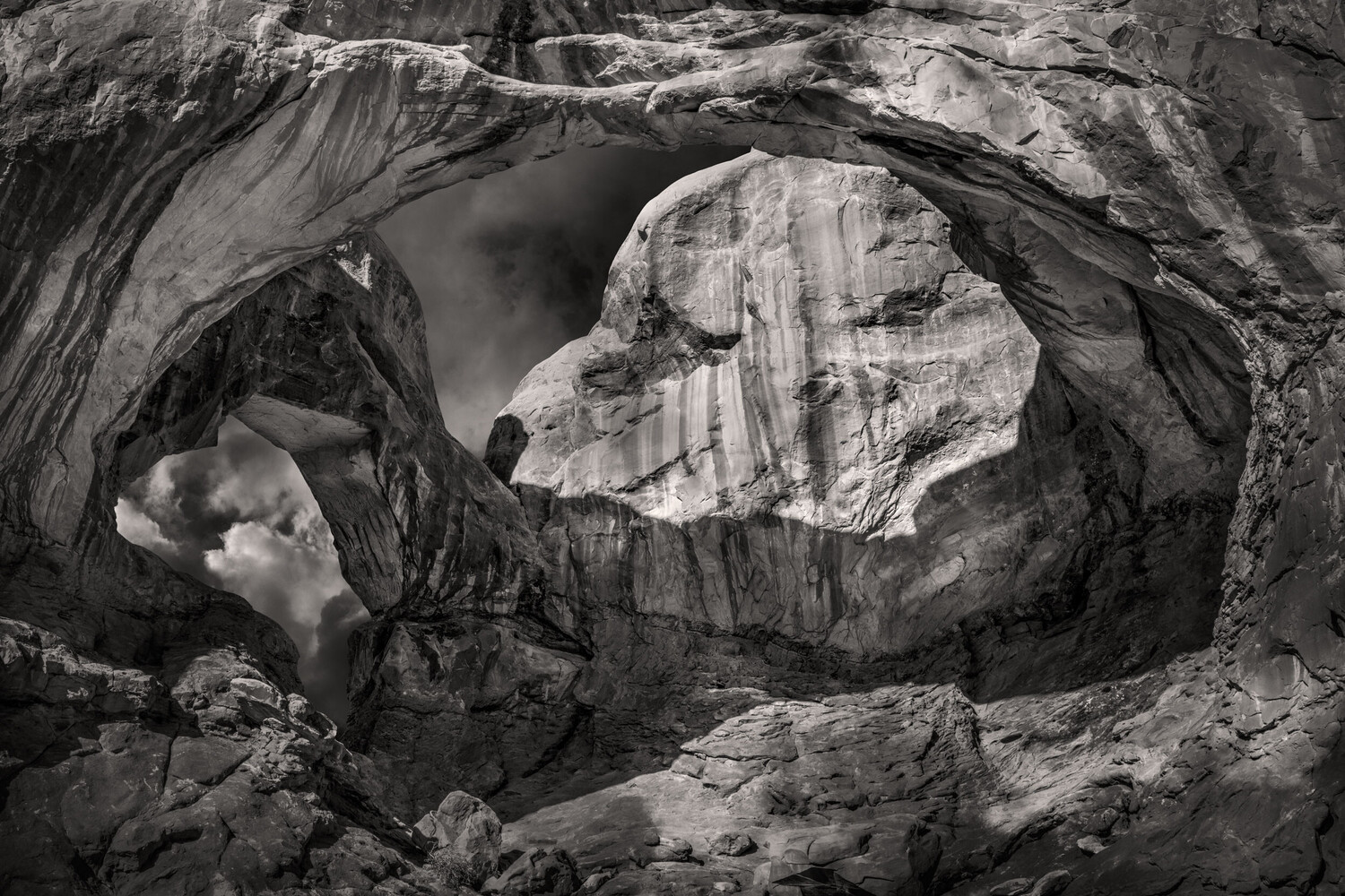 Black and white view through a sandstone arch, with a smaller opening framing clouds and a rock fin lit by sunlight at center.
